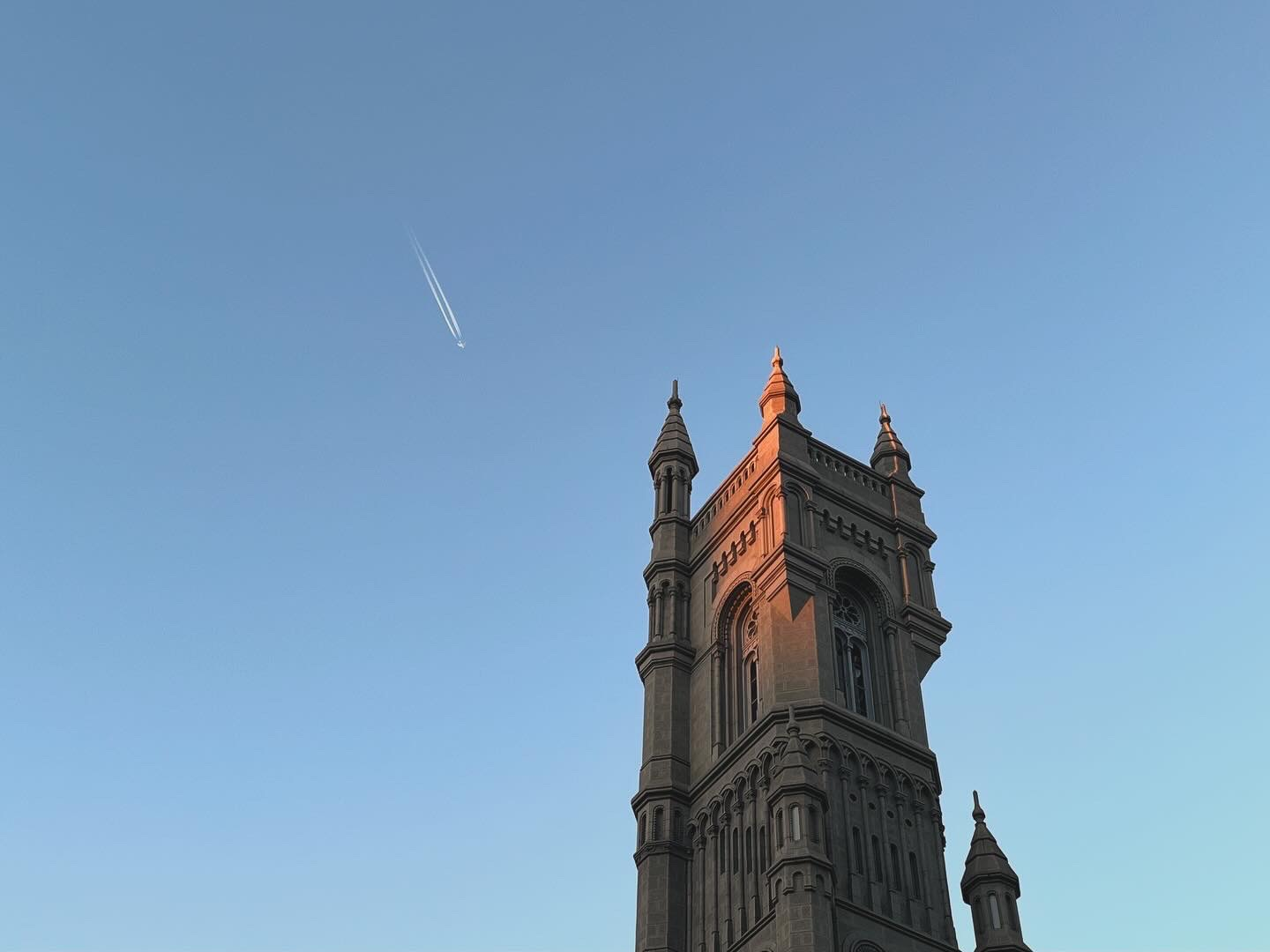 Sunlit building tower beneath a blue sky with an airplane and its contrails.